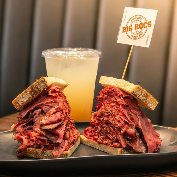 Two towering pastrami sandwiches on rye bread with a drink and a "Big Rocs" flag.