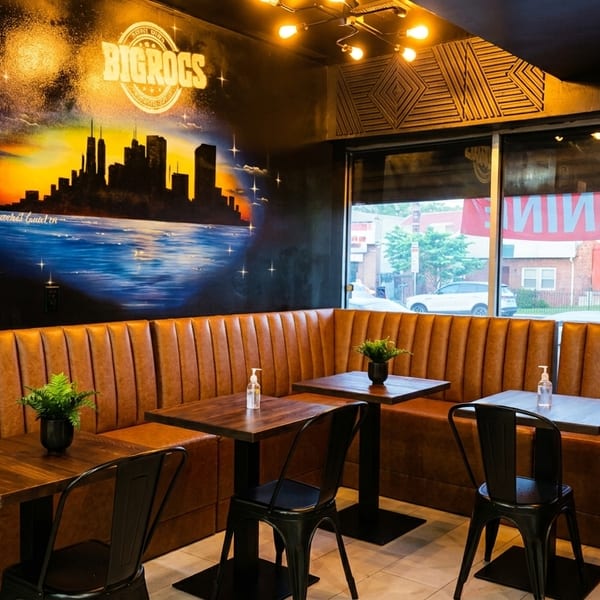 Restaurant interior with brown booth seating, dark tables, black chairs, and a cityscape mural on the wall.