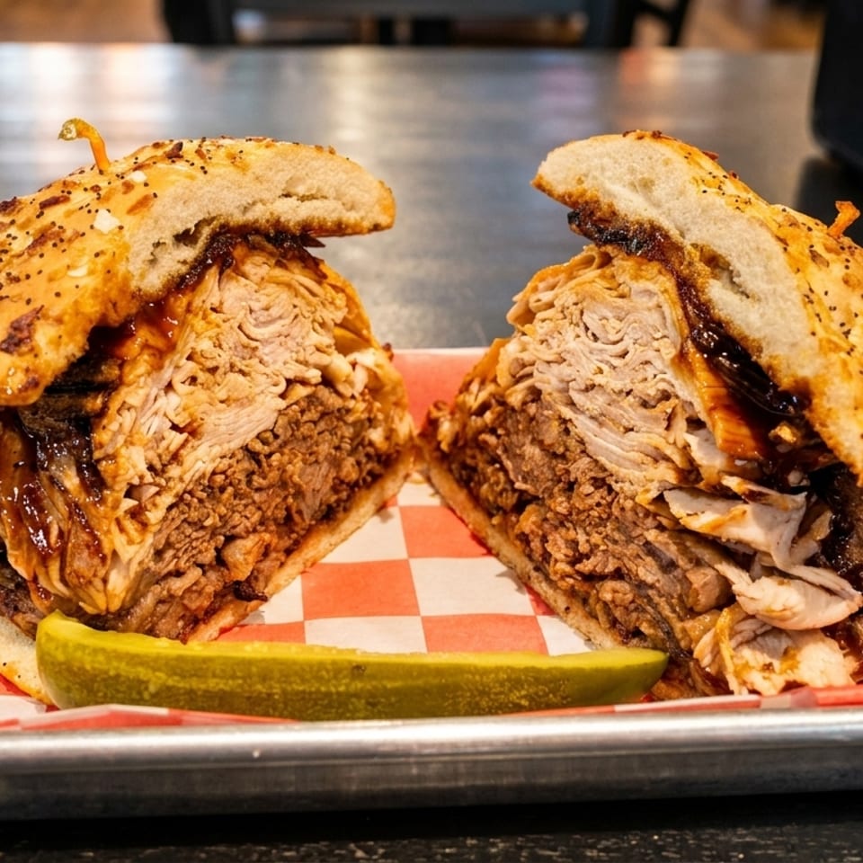 A large, sliced BBQ sandwich with pulled pork and brisket, served with a pickle spear on a tray.